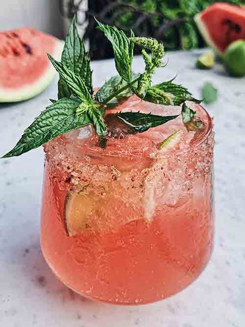 A Gin and Tonic infused with Watermelon and Mint in a glass served over ice and garnished with sugar mixture around the rim